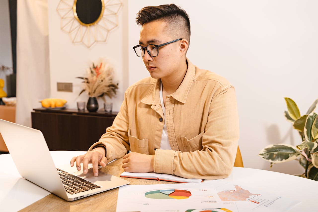 Young Asian man focused on work, using a laptop at a modern home office.