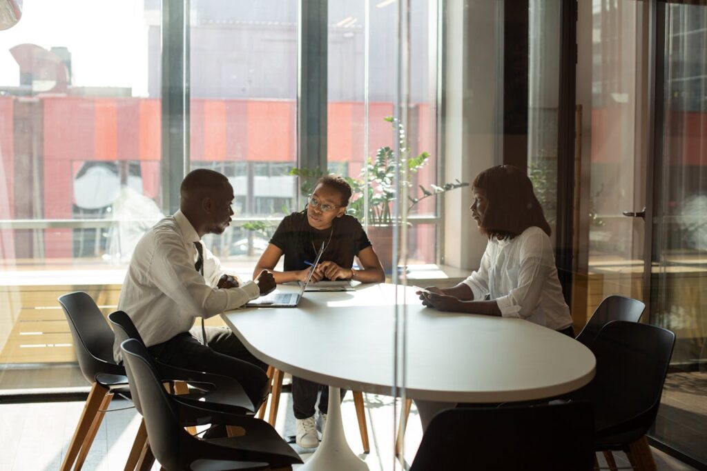 Three colleagues have a discussion around a table in a modern, glass-walled office.